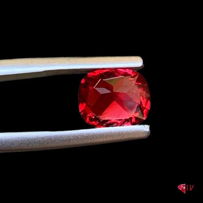May include: A faceted, cushion-cut, red gemstone is held by tweezers against a black background. The gem has a brilliant, reflective surface, showcasing its internal structure and color. The tweezers are silver.