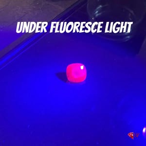 May include: A bright pink, square-shaped gemstone illuminated under a blue fluorescent light. The text "UNDER FLUORESCE LIGHT" is displayed in white, angled across the top of the image. The gemstone appears to glow.