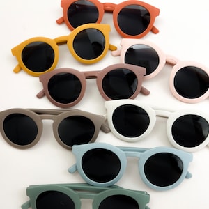 Puede incluir: Una colección de gafas de sol con montura redonda en varios colores: naranja, amarillo, rosa, marrón, gris, blanco, azul y verde. Cada par tiene lentes oscuras. Las gafas de sol están dispuestas sobre una superficie blanca.