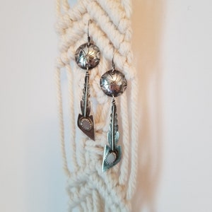May include: A pair of silver earrings with a feather design and a round, textured silver charm. The earrings are hanging from a white macrame wall hanging.