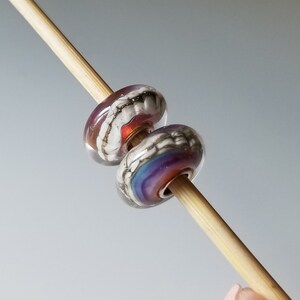 Peut inclure: Deux perles en verre avec des motifs complexes. La perle du haut est blanche avec un tourbillon argenté et un accent rouge. La perle du bas est transparente avec un tourbillon violet, bleu et rose.