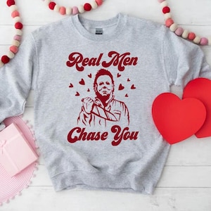 Op de afbeelding: Een grijs sweatshirt met een rode afbeelding van een gemaskerd figuur dat een mes vasthoudt. De afbeelding zegt "Real Men Chase You" met harten rond de figuur.