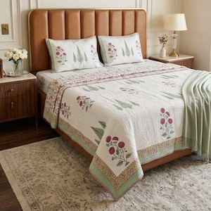 May include: A white quilted bedspread and two matching pillow shams featuring a repeating pattern of pink floral motifs and green leaf designs. The bedding has a light green border with a small floral print and is draped over a brown leather headboard.