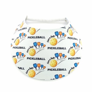 Op de afbeelding: Witte visor met een herhalend patroon van pickleball-rackets, ballen en de tekst "Have a Ball!" en "Pickleball".
