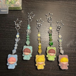 May include: Five keychains with star-shaped clasps and beaded strands. Each keychain features a small, plush figure in different colors: blue, green, yellow, and pink. The figures have closed eyes and brown or yellow hair.