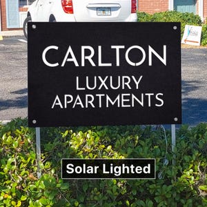 May include: A rectangular black sign with white text reading CARLTON LUXURY APARTMENTS. The sign features four silver corner grommets and is mounted on two metal stakes. A separate black rectangular label below the sign reads Solar Lighted.