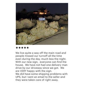 May include: A night-time view of a house with a lit-up address sign that reads "2291". The sign is mounted on a post in front of a house with a dark, wooden exterior. The house is surrounded by bushes and trees.
