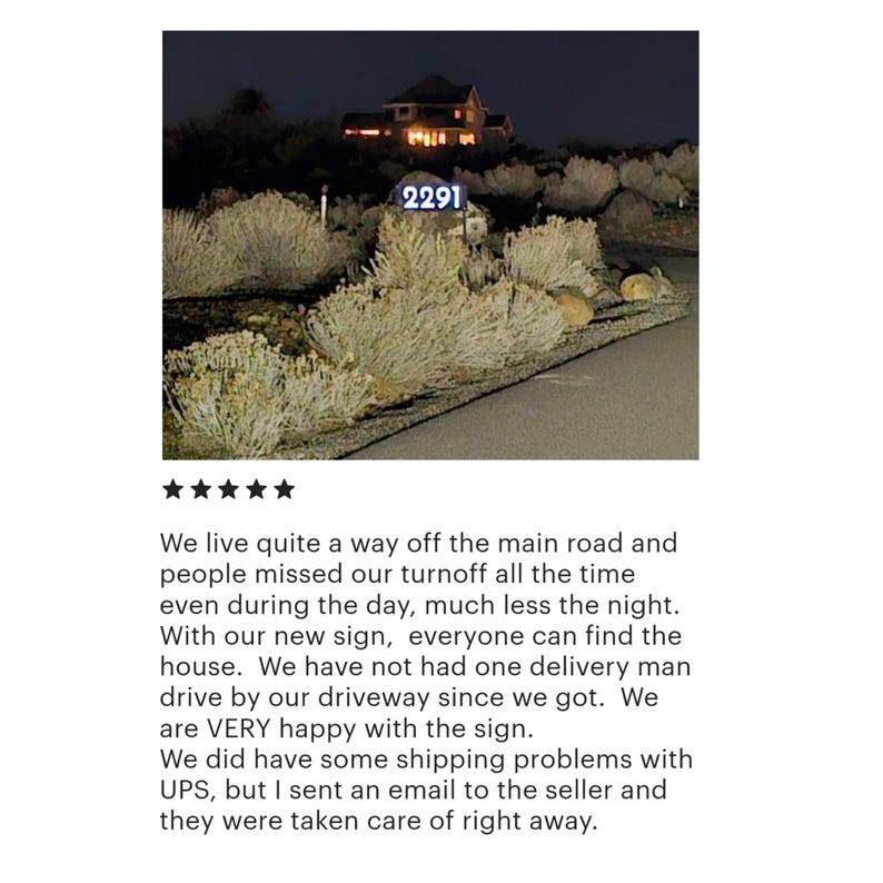 May include: A night-time view of a house with a lit-up address sign that reads "2291". The sign is mounted on a post in front of a house with a dark, wooden exterior. The house is surrounded by bushes and trees.