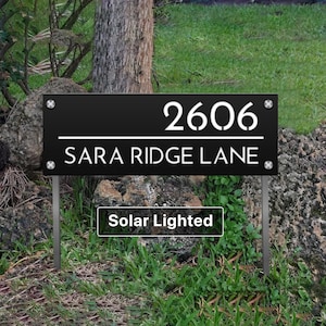 May include: A rectangular black address sign featuring the white text 2606 and SARA RIDGE LANE separated by a horizontal line. The sign is secured with four silver corner screws and mounted on two metal stakes, with a Solar Lighted label below.