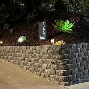 May include: A black metal house number sign with white numbers, reading "1772", and the words "Paso Viento" below. The sign is mounted on a wooden post and is surrounded by green plants and a stone wall.