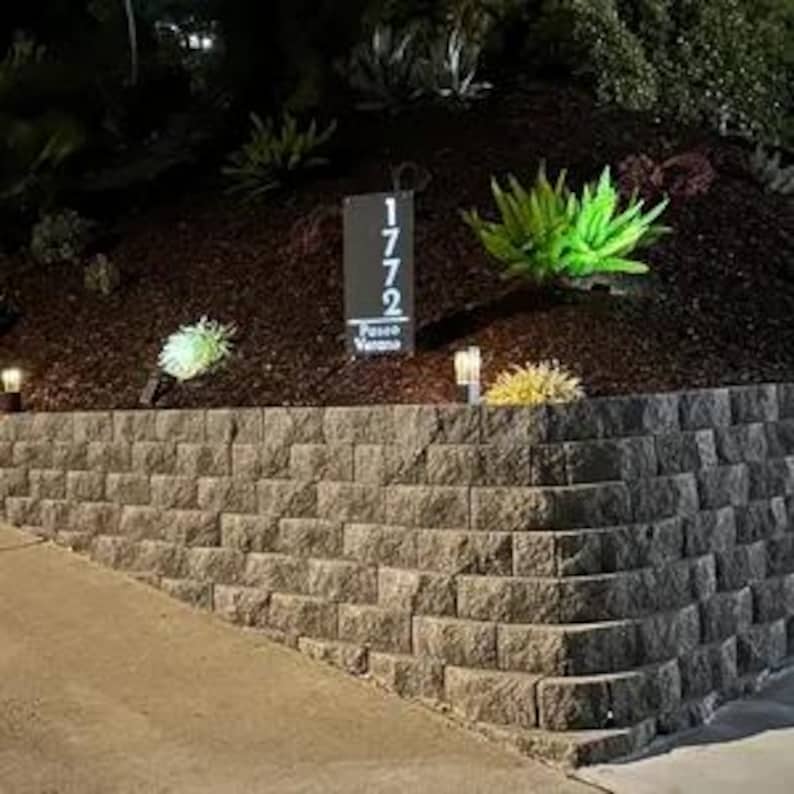 May include: A black metal house number sign with white numbers, reading "1772", and the words "Paso Viento" below. The sign is mounted on a wooden post and is surrounded by green plants and a stone wall.