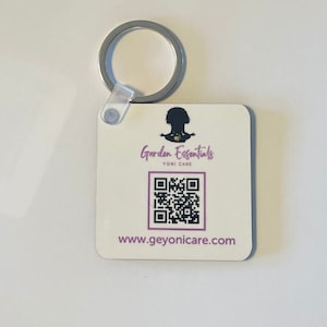 May include: White square keychain with a black silhouette of a person sitting in a yoga pose. The text "Garden Essentials Yoni Care" is printed in purple. A QR code is printed below the text. The text "www.geyonicare.com" is printed below the QR code.