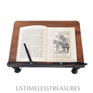 May include: A wooden book stand with a book open to a page with a black and white illustration of horses and a person. The stand has a dark brown finish and is supported by four small, rounded feet.