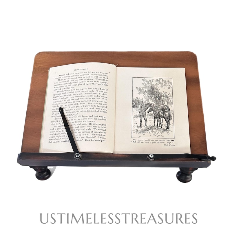 May include: A wooden book stand with a book open to a page with a black and white illustration of horses and a person. The stand has a dark brown finish and is supported by four small, rounded feet.