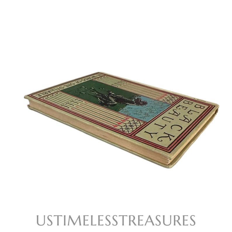 May include: An antique book titled "Black Beauty" with a green, red, and cream cover. The cover features an illustration of a horse and rider. The book is slightly angled, showing the spine and edges.