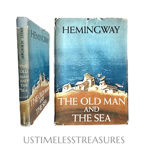May include: A vintage hardcover book titled The Old Man and the Sea by Ernest Hemingway, featuring a blue and brown illustrated dust jacket depicting a coastal village scene. The book is shown from both the front cover and the spine perspective.