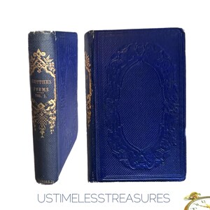 May include: Two antique books with blue covers. One book is standing upright, displaying gold lettering and ornate designs. The other book is lying flat, showcasing a gold-framed embossed design. The books are from the "Whittier's Poems" collection.