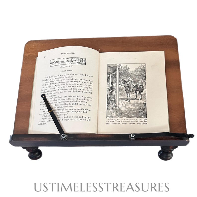 May include: A dark wooden book stand with a book open to Chapter V of "Black Beauty". The book has black and white illustrations and text. The stand has a small shelf and is supported by four rounded feet. The text "USTIMELESSTREASURES" is at the bottom.