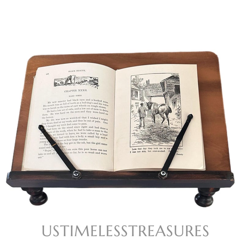 May include: A wooden book stand with a dark brown finish, holding an open book. The book has black and white illustrations and text. The stand has a sloped top and a lower shelf with decorative feet. The text on the book reads "BLACK BEAUTY".