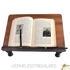 May include: A dark wooden book stand with a book open to a page with black and white images and text. The stand has a lip to hold the book and a strap to keep the book open. The text "USTIMELESSTREASURES" is at the bottom.