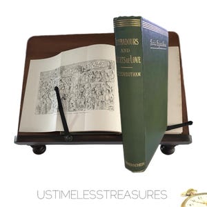 May include: A green hardcover book titled "Troubadours and Courts of Love" by J.R. Rowbotham, resting on a wooden book stand with a black metal page holder. The book is open to a page with a black and white illustration.