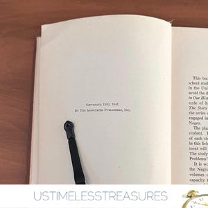 May include: Open book with text, copyright 1935, 1942, by The Associated Publishers, Inc. A black ribbon bookmark is inserted between the pages. The book is on a wooden surface.