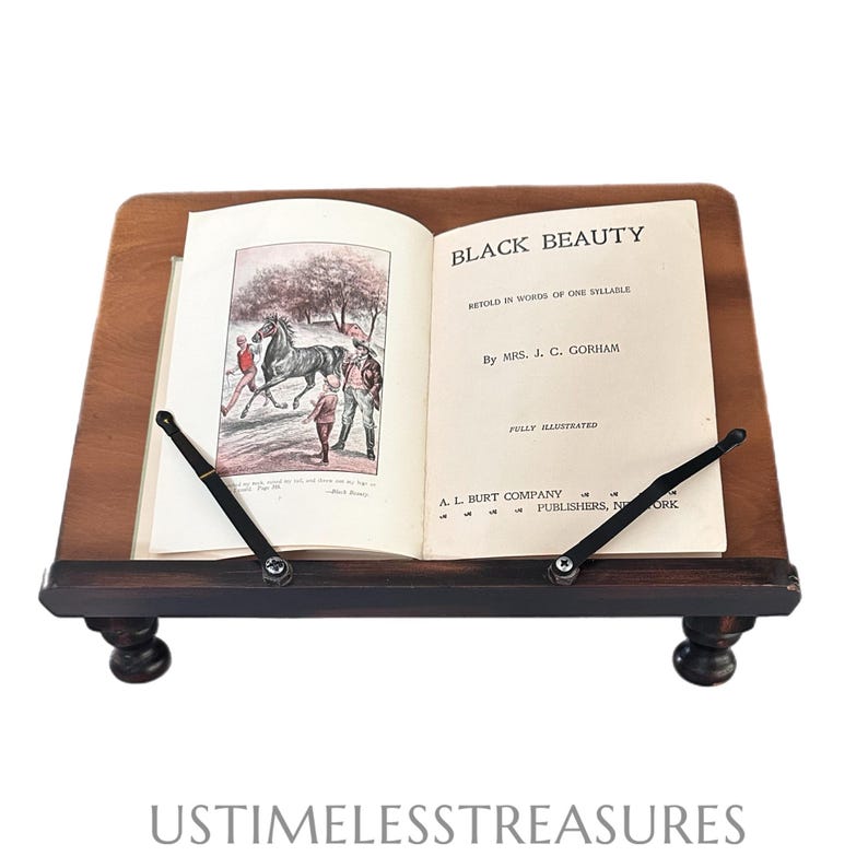 May include: A wooden book stand with a book open to the title page of "Black Beauty." The book is held in place by black metal supports. The stand has a dark brown finish and rests on four small, rounded feet. The text "USTIMELESSTREASURES" is at the bottom.