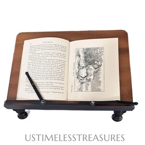 May include: A wooden book stand with a dark brown base and a lighter brown top. The stand holds an open book with text and an illustration. The stand has two small, round feet and a metal arm to hold the book open.