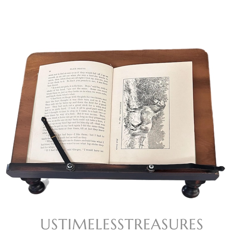 May include: A wooden book stand with a dark brown base and a lighter brown top. The stand holds an open book with text and an illustration. The stand has two small, round feet and a metal arm to hold the book open.