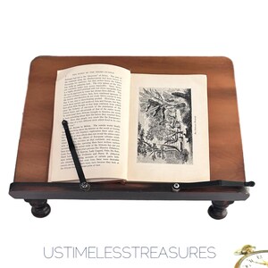 May include: A wooden book stand with a dark brown base and a light brown top. An open book is held in place by a black metal arm. The book has black and white illustrations and text. The text "USTIMELESSTREASURES" is at the bottom.