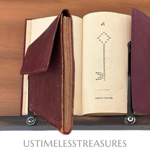 May include: An open antique book with a maroon leather cover, held upright by black metal bookends. The page displays a stylized letter "A" and the words "Fourth Edition."