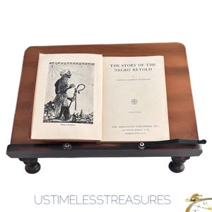 May include: An open book titled "The Story of the Negro Retold" by Carter Godwin Woodson, resting on a wooden book stand. The book features a black and white illustration and text. The stand is dark brown with decorative feet.