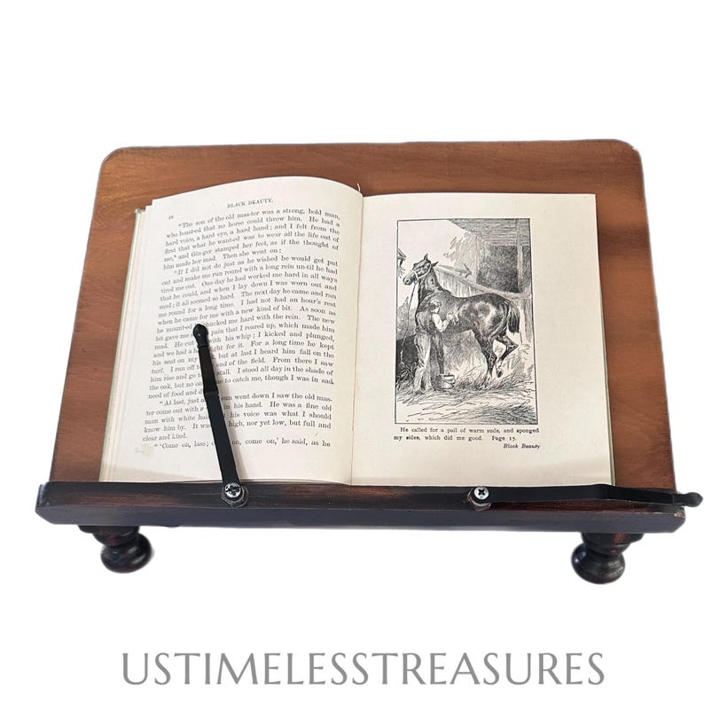May include: A wooden book stand with a book open to a page with a horse illustration. The stand is dark brown with a slanted top and a lower shelf. The book is held in place by a black metal arm. The text "Black Beauty" is visible.
