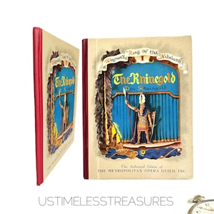 May include: A vintage book titled "The Rhinegold" with a red spine and a colorful illustration of a character on a stage. The book is part of Wagner's "Ring of the Nibelung" series and is an authorized edition of the Metropolitan Opera Guild.
