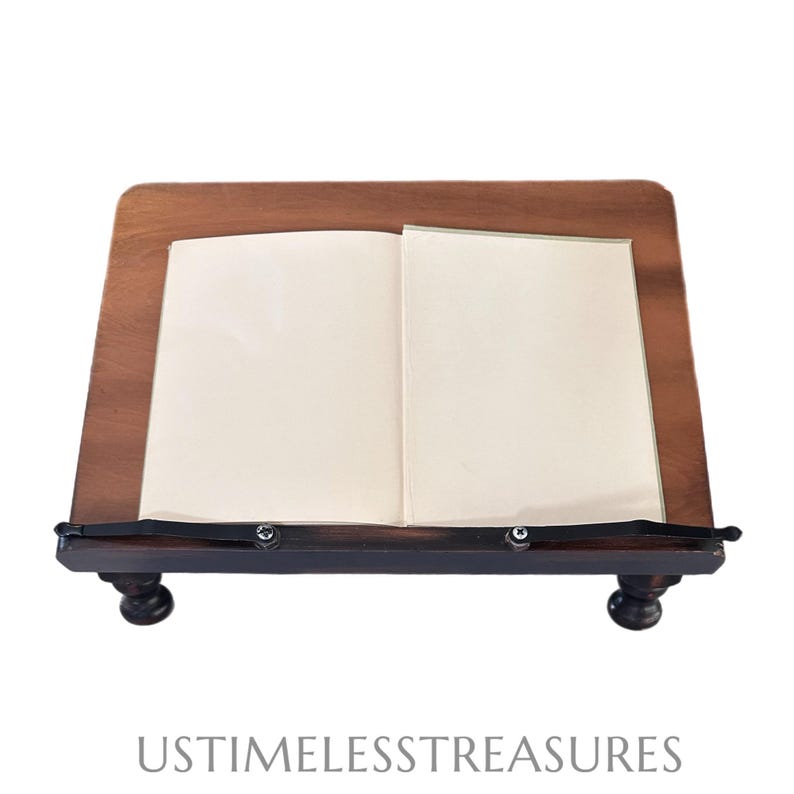 May include: A wooden book stand with an open book. The stand is made of dark brown wood with a rectangular top and two small, round feet. The open book has off-white pages. The text "USTIMELESSTREASURES" is at the bottom.