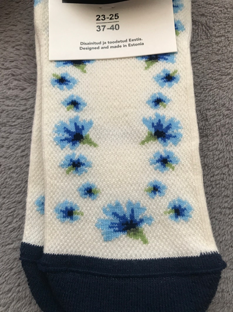 May include: A pair of white socks with a blue floral pattern. The socks have a navy blue cuff and the text "23-25 37-40 Disainitud ja tootedud Eestis. Designed and made in Estonia" is printed on the tag.