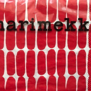 May include: A close-up of a red and white patterned napkin with the word "marimekko" in black block letters. The napkin has a repeating pattern of red oval shapes on a white background. The napkin is made of a textured paper material.