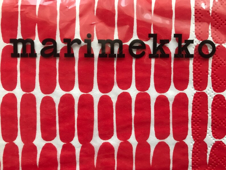 May include: A close-up of a red and white patterned napkin with the word "marimekko" in black block letters. The napkin has a repeating pattern of red oval shapes on a white background. The napkin is made of a textured paper material.