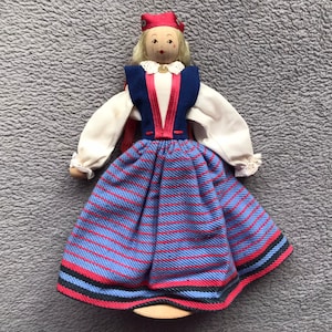 May include: A wooden doll wearing a blue and red striped skirt, a white blouse, a blue vest, and a red hat. The doll has blonde hair and a painted face.