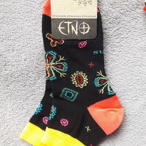 May include: A pair of black ankle socks with a colorful geometric pattern. The socks have a yellow cuff and an orange heel. The socks have a tag attached that says "ETNO" and "36-38".