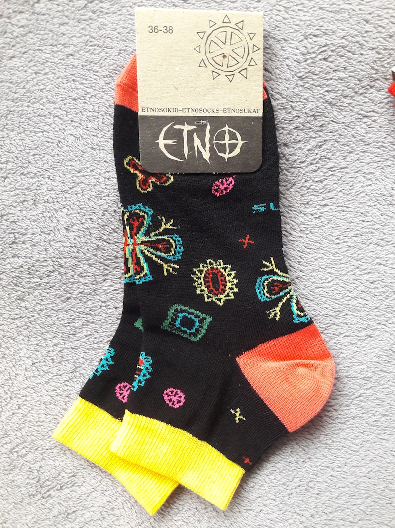 May include: A pair of black ankle socks with a colorful geometric pattern. The socks have a yellow cuff and an orange heel. The socks have a tag attached that says "ETNO" and "36-38".