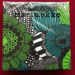 May include: A package of Marimekko napkins with a vibrant floral design in green, teal, black, and white. The napkins feature bold, abstract flower patterns and the Marimekko logo in black. The background is a solid red.