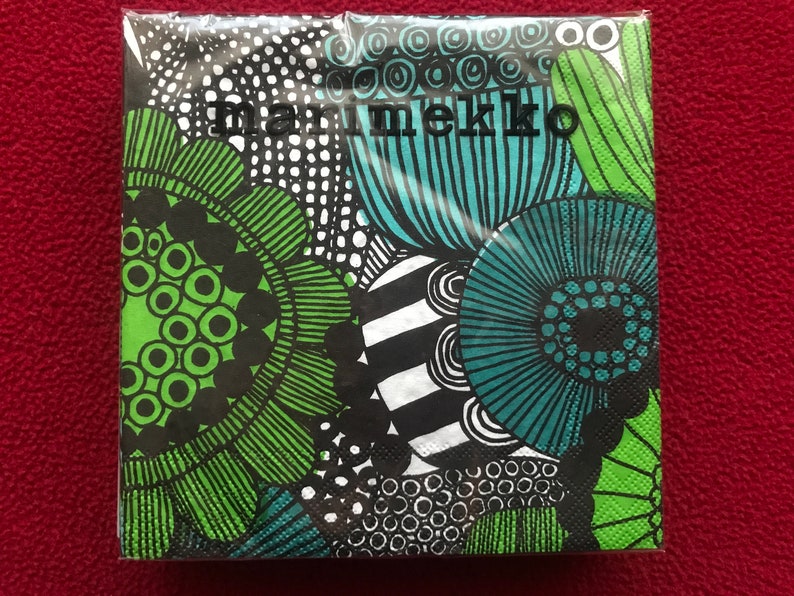 May include: A package of Marimekko napkins with a vibrant floral design in green, teal, black, and white. The napkins feature bold, abstract flower patterns and the Marimekko logo in black. The background is a solid red.