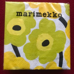 May include: A package of Marimekko paper napkins featuring a bold floral pattern. The design includes large, stylized flowers in shades of green and yellow against a white background. The brand name "marimekko" is printed in black.