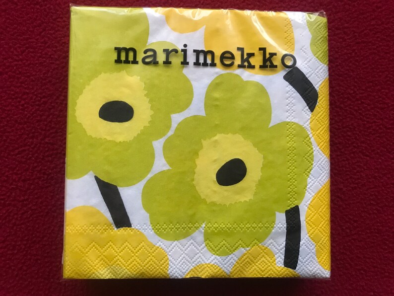 May include: A package of Marimekko paper napkins featuring a bold floral pattern. The design includes large, stylized flowers in shades of green and yellow against a white background. The brand name "marimekko" is printed in black.