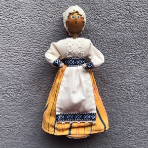 May include: A wooden doll wearing a white dress with yellow and blue embroidery. The doll has a white head with painted features and a white cap.