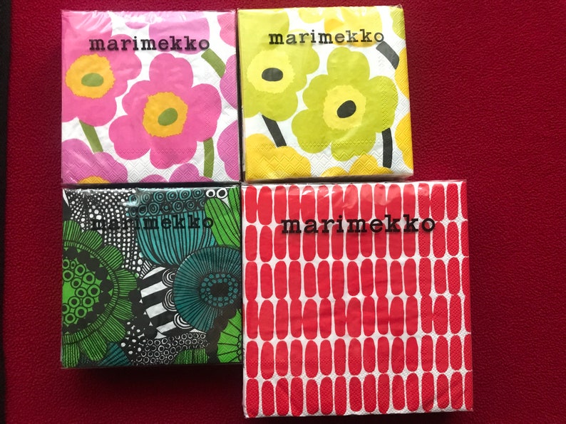 May include: Four packages of Marimekko brand paper napkins with floral and abstract patterns. The napkins feature designs in pink, yellow, green, and red, with the brand name printed in black.