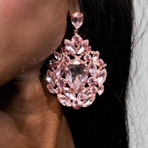 May include: A large, pink crystal earring with a teardrop-shaped top and a cluster of pink crystals in a floral design. The earring is rose gold in color and has a large, central crystal.