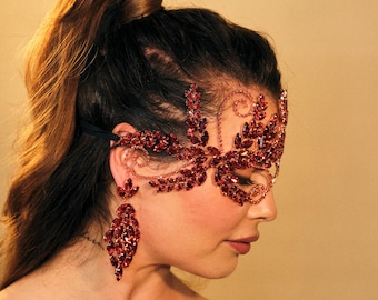 May include: A red jeweled masquerade mask with a delicate floral design. The mask is worn by a person with their hair pulled back in a ponytail.