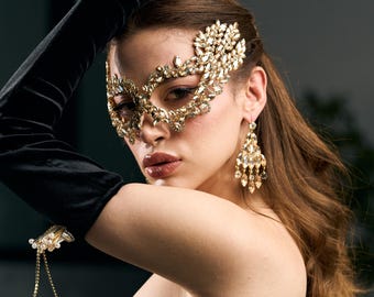 May include: A glamorous gold-coloured masquerade mask embellished with crystals. The mask covers the eyes and forehead, paired with matching chandelier earrings. The person is wearing long black velvet gloves and a black strapless top, creating an elegant look.
